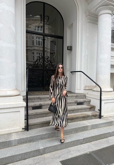 Mujer con un vestido largo de estampado de cebra y gafas de sol baja unas escaleras de piedra frente a una puerta alta y negra con el número 12 encima.