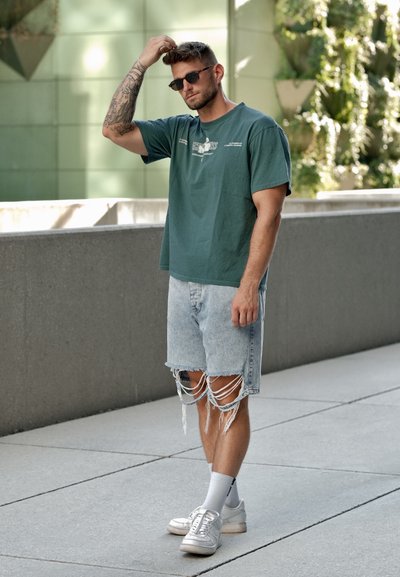 Camiseta de algodón verde con un estampado gráfico, combinada con pantalones cortos de mezclilla clara desgastados. Zapatillas blancas y calcetines de tobillo completan el look.