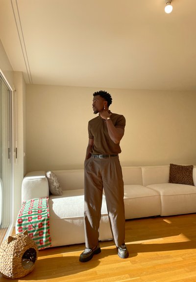 Hombre con camisa y pantalones marrones de pie cerca de un sofá blanco con manta estampada y cesta para gatos en una sala de estar iluminada por el sol.