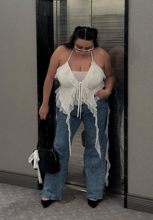 Top blanco con volantes en capas, jeans de talle alto azul claro con adornos y tacones negros de punta. Bolso negro con un detalle de cinta.
