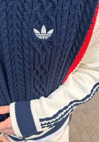 Pull en maille bleu marine avec logo Adidas blanc, manches blanches avec rayures bleu marine, et une bordure rouge sur le côté droit, porté par une personne croisant les bras.