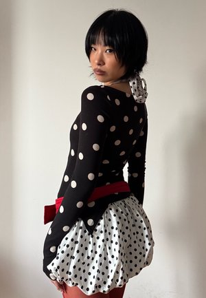 Persona con cabello corto y negro que lleva una blusa y falda de lunares blancos y negros con cinturón rojo, mirando por encima del hombro contra una pared lisa.