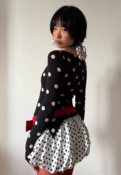 Persona con cabello corto y negro que lleva una blusa y falda de lunares blancos y negros con cinturón rojo, mirando por encima del hombro contra una pared lisa.