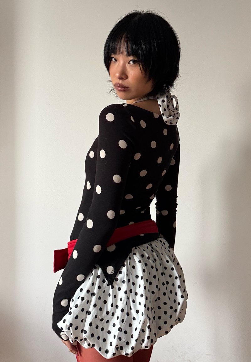 Persona con capelli corti neri che indossa un top e una gonna a pois bianchi e neri con cintura rossa, che guarda oltre la spalla contro una parete uniforme.
