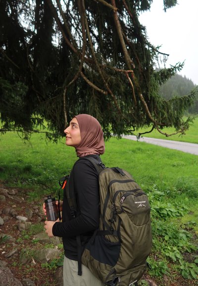 Mochila de senderismo verde con correas acolchadas, sosteniendo una botella de agua, de pie junto a un árbol, vistiendo una camiseta de manga larga negra y un pañuelo marrón en la cabeza.