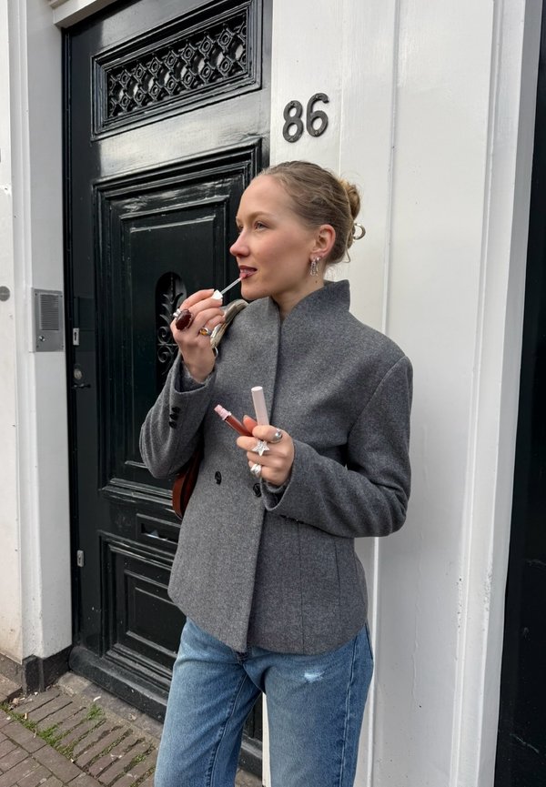 Vrouw met licht haar in een grijze blazer en blauwe spijkerbroek brengt lipgloss aan buiten een zwarte deur met nummer 86.