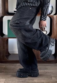 Personne portant un pantalon large noir et des bottes noires épaisses avec des détails métalliques, posant avec une jambe pliée à l'intérieur sur un sol en bois.