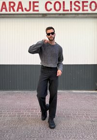 Pulover gri cu dungă, blugi negri cu talie înaltă și adidași negri; pozat lângă un perete alb și gri cu litere roșii deasupra.