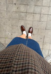 Personne portant des mocassins marron, des chaussettes blanches, une jupe en denim bleue et un haut à carreaux marron debout sur un trottoir carrelé.