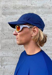 Blå caps, hvite solbriller med oransje linser, teksturert blå skjorte. Profilbilde mot en teksturert betongbakgrunn.