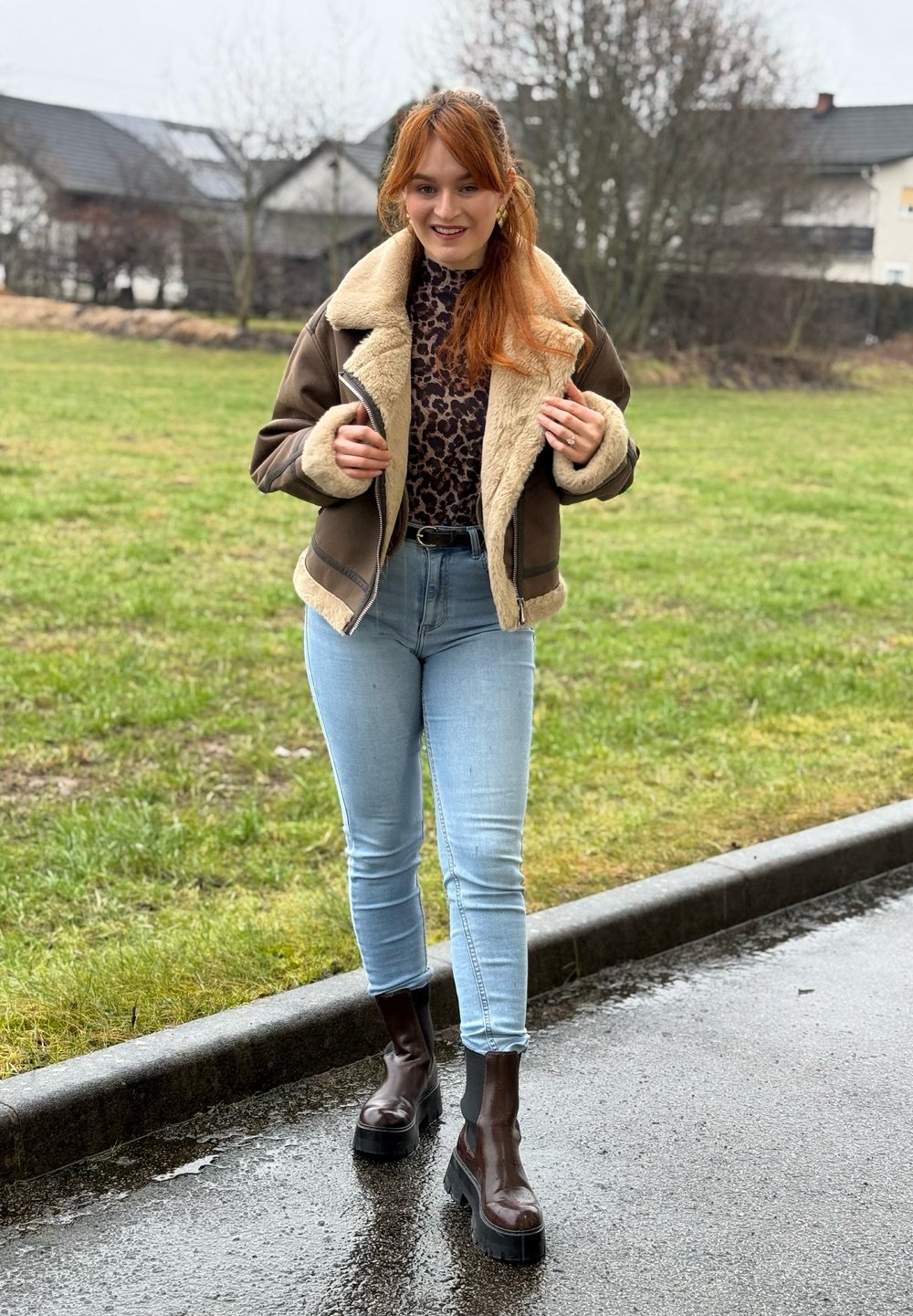 Jeune femme aux cheveux roux portant une veste en peau retournée marron, un haut imprimé léopard, un jean bleu clair et des bottes marron foncé sur un trottoir humide.