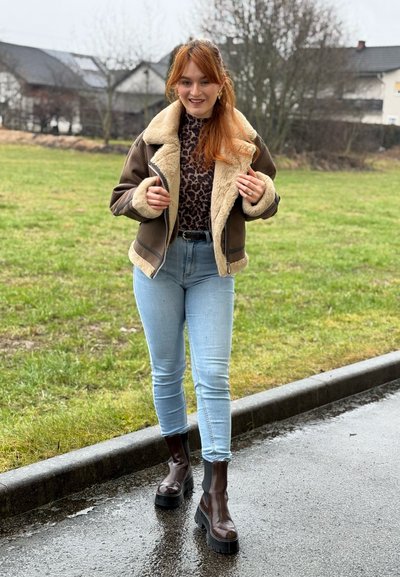 Mujer joven con cabello rojo, vestida con chaqueta marrón de piel, blusa con estampado de leopardo, jeans azul claro y botas marrón oscuro sobre un pavimento mojado.