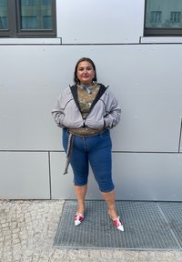 Světle šedá bunda s kapucí přes olivově zelený vzorovaný top, modré krátké džíny a červenobílé boty, pózující před šedou zdí.