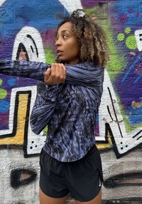 Črna in vijolična dolga majica za vadbo z valovitimi vzorci, v kombinaciji s črnimi kratkimi hlačami. Model se razteguje ob barvito grafitno ozadje.