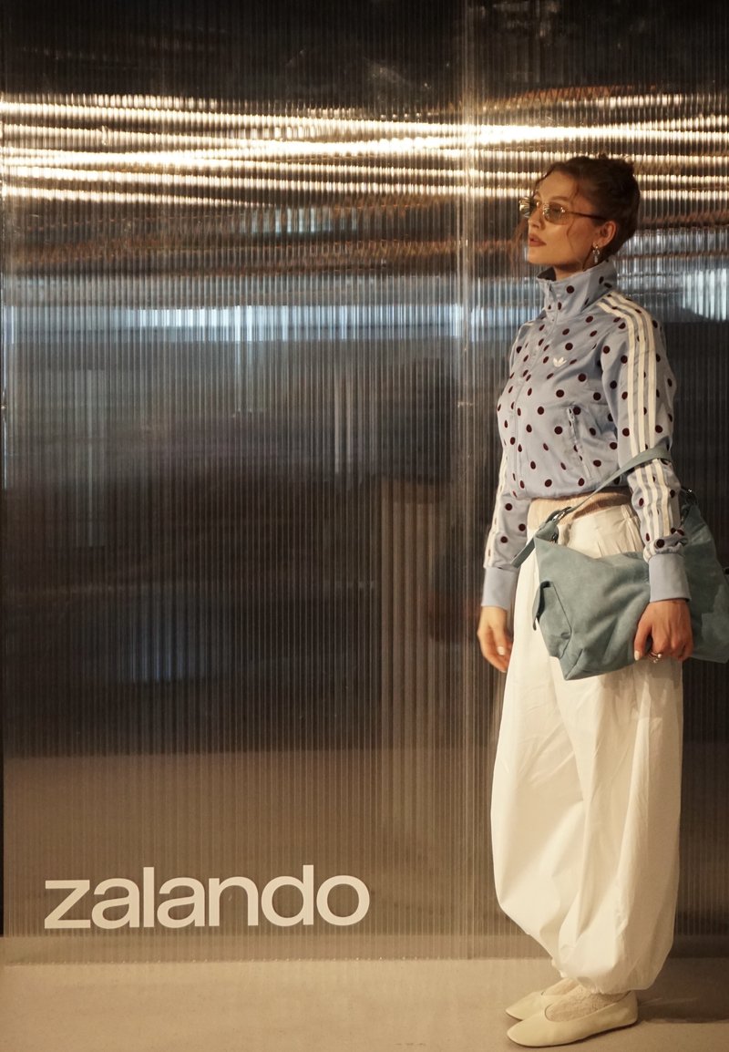 Donna con giacca a pois, pantaloni bianchi larghi, scarpe beige, tiene una borsa blu, in piedi accanto a una parete trasparente e strutturata con la scritta "zalando".