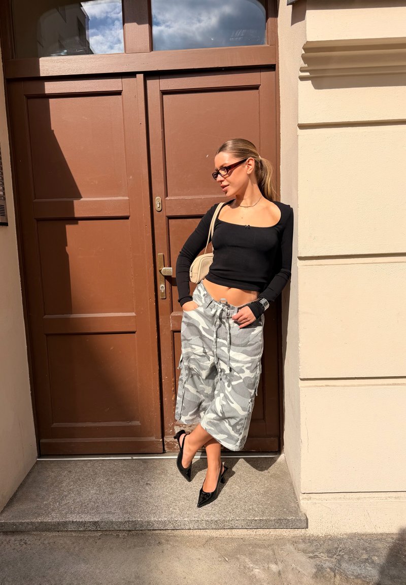 Femme en crop top noir à manches longues, pantalon camouflage à jambes larges, et talons noirs appuyée contre un mur crème au soleil près d'une porte marron fermée.
