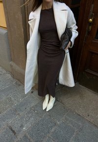 Femme portant une robe ajustée brun foncé, un manteau blanc, des bottes blanches à bout pointu, tenant une pochette noire, debout sur un pavé en pierre près d'une porte en bois.