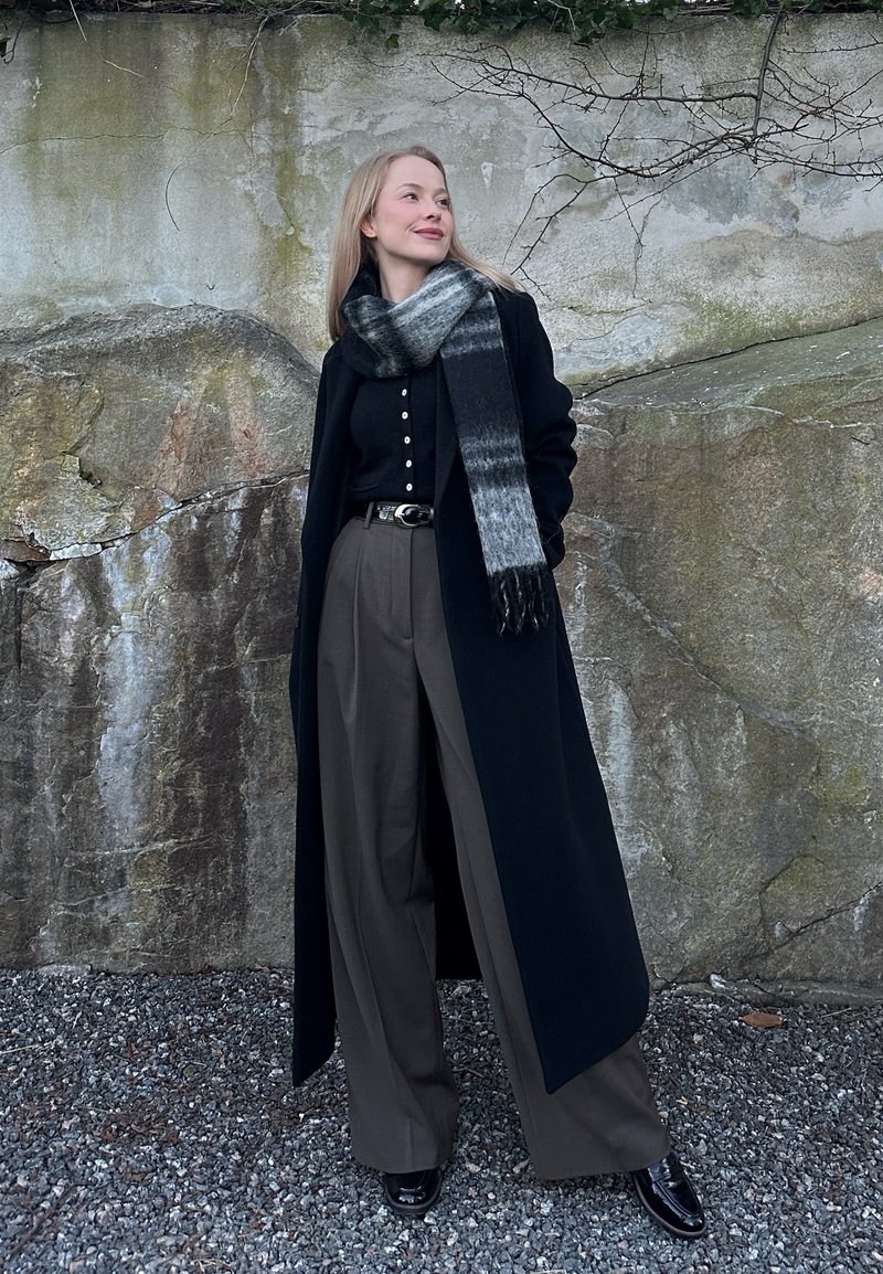 Νεαρή γυναίκα στέκεται σε χαλίκι μπροστά από έναν πέτρινο τοίχο, φορώντας ένα μακρύ μαύρο παλτό, γκρι φαρδιά παντελόνια, μαύρο τοπ, μαύρα παπούτσια και γκρι κασκόλ.