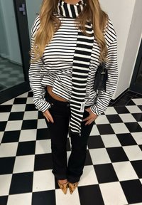 Črno-beli črtasti pulover z visokim ovratnikom, kombiniran s črnimi hlačami in rjavimi petami s potiskom, poudarjen z ujemajočim se črtastim šalčkom.