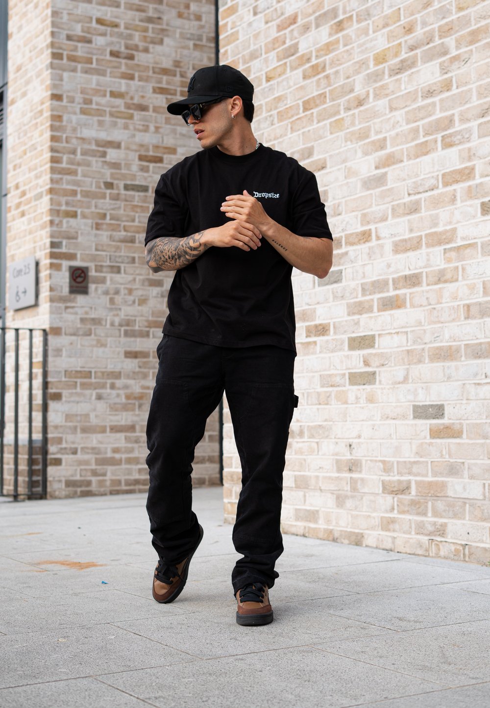 Black oversized t-shirt featuring the "Dropsize" logo, paired with black cargo trousers and brown trainers. Sunglasses and a black cap complete the outfit.