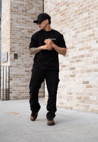 Black oversized t-shirt featuring the "Dropsize" logo, paired with black cargo trousers and brown trainers. Sunglasses and a black cap complete the outfit.