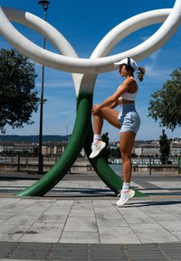 Atleta em roupa desportiva branca e calções cinzentos, fazendo alongamentos ao lado de uma escultura moderna abstrata verde e branca numa praça. Céu azul limpo.