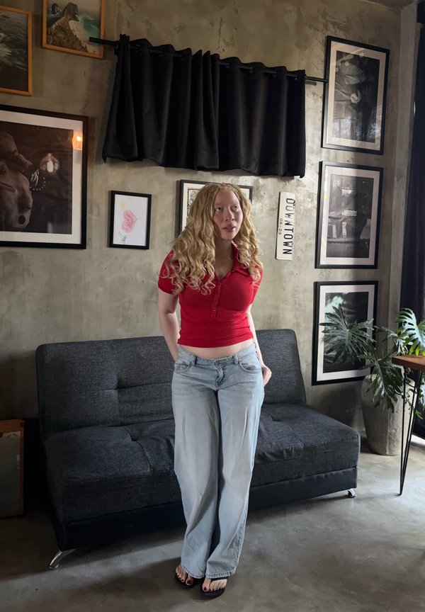 Femme aux longs cheveux bouclés portant une chemise rouge et un jean bleu clair debout dans un salon avec un canapé sombre, des tableaux encadrés et une plante en pot.