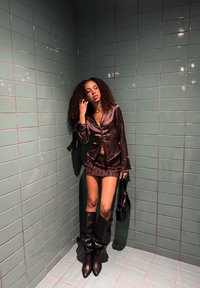 Braunes Satin-Hemd zum Knöpfen, schwarzer Minirock mit Pailletten, kniehohe schwarze Stiefel und eine schwarze Handtasche vor einer grünen Fliesenwand.