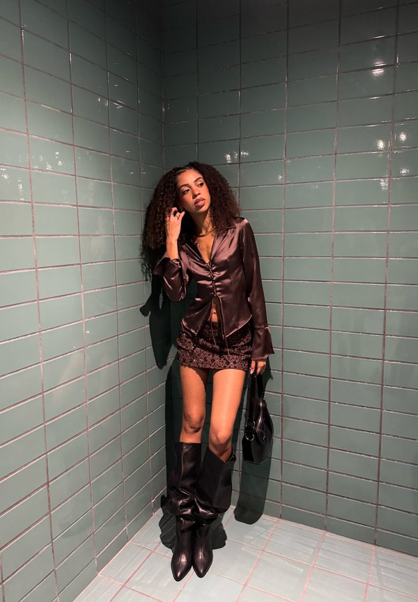 Brun satin skjorta med knappar, svart paljettminikjol, svart knästövlar och en svart handväska mot en grön kakelvägg.