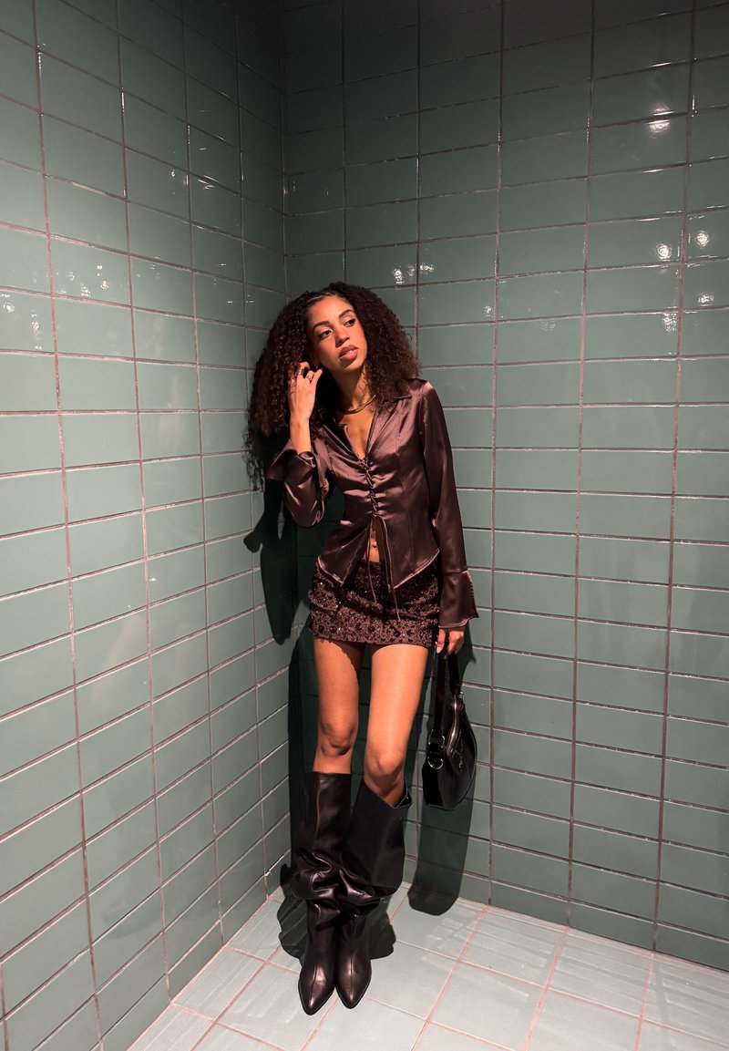 Camicia marrone in satin con bottoni, minigonna nera con paillettes, stivali neri al ginocchio e borsa nera davanti a una parete piastrellata verde.