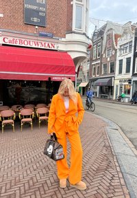 Costume orange ajusté avec un pantalon à jambes larges et une taille ceinturée, associé à des chaussures à plateforme beige. Tient un sac noir à motifs avec un texte graphique.