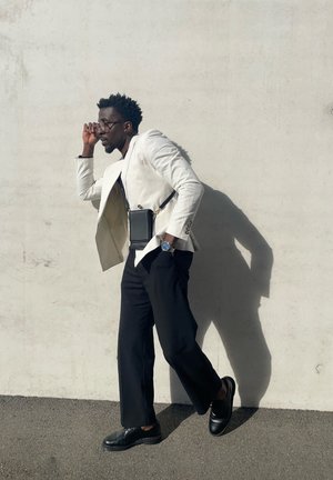 Blazer blanco de lino, pantalones anchos negros, bolso negro con correa, zapatos negros, accesorios con gafas y un reloj. Fondo neutro.