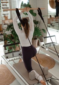 Nainen polvillaan Pilates-reformerissa, pitäen kädessään puista tankoa ylhäällä kiinnitettynä hihnoihin kirkkaassa kuntosalissa, jossa on kasveja ja peilejä.