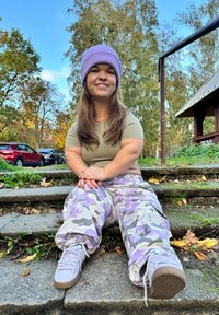 Casualna odjeća koja se sastoji od uske maslinasto zelene majice i širokih lavandastih kamuflažnih hlača. Dodatno su tu lavandaste tenisice. Vanjsko okruženje s bogatom zelenilom.