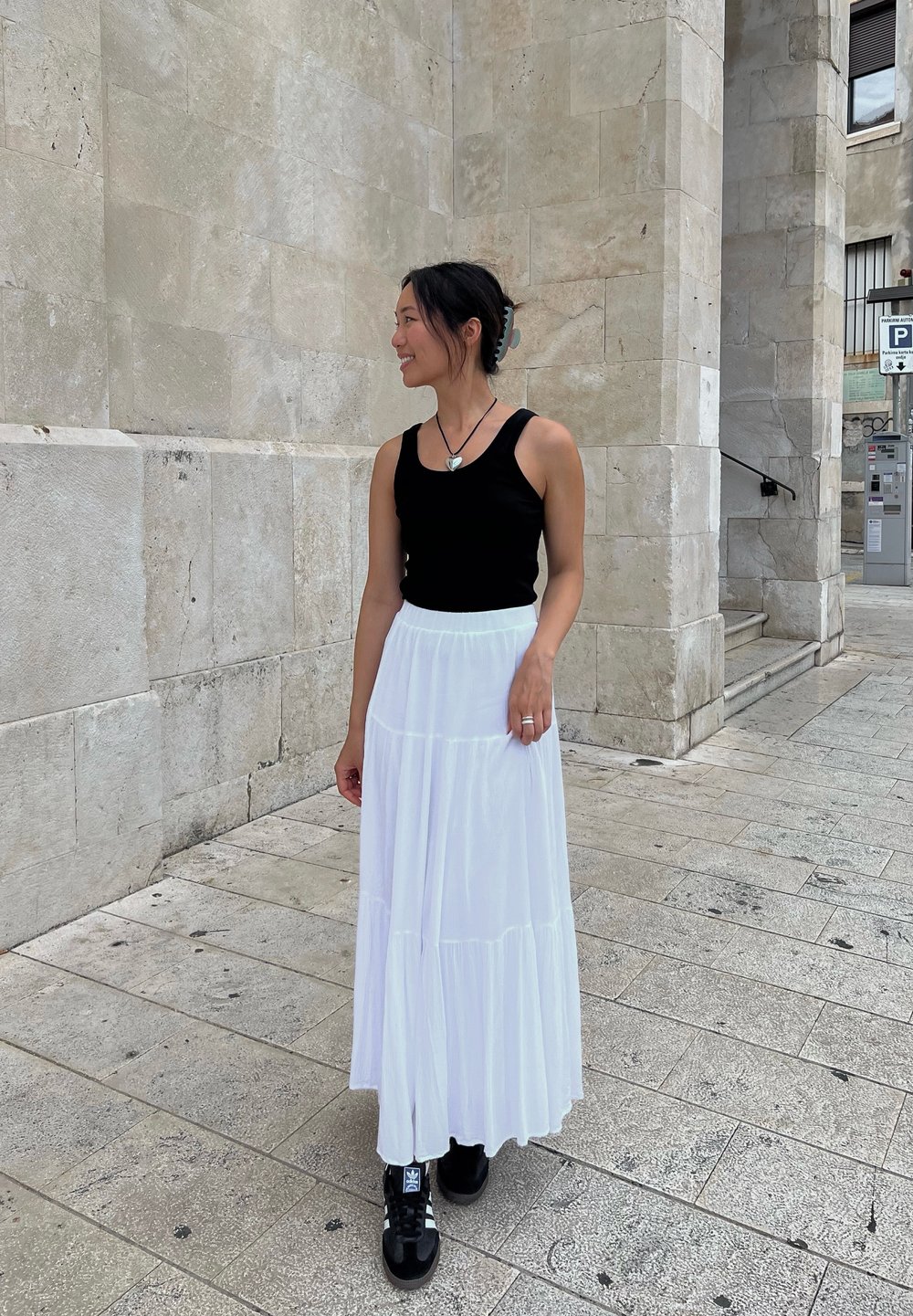 Μαύρο αμάνικο μπλουζάκι, λευκή φούστα μάξι με επίπεδα, μαύρα αθλητικά παπούτσια με λευκές ρίγες, ασημένιο δαχτυλίδι, στέκεται σε κυβόλιθους μπροστά από ένα φωτεινό τοίχο.