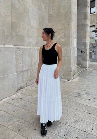 Μαύρο αμάνικο μπλουζάκι, λευκή φούστα μάξι με επίπεδα, μαύρα αθλητικά παπούτσια με λευκές ρίγες, ασημένιο δαχτυλίδι, στέκεται σε κυβόλιθους μπροστά από ένα φωτεινό τοίχο.