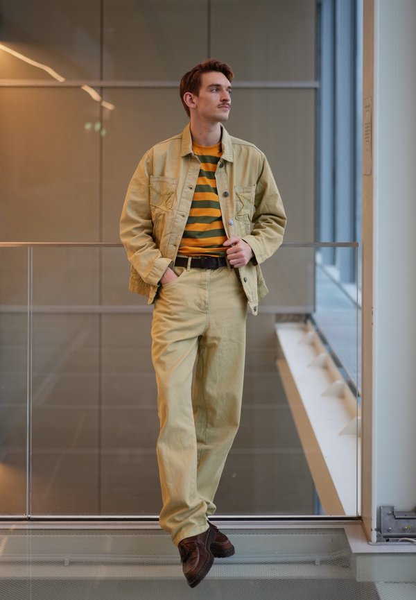 Jeune homme en veste et pantalon beige avec une chemise rayée, debout à l'intérieur près d'une rambarde en verre, regardant sur le côté.