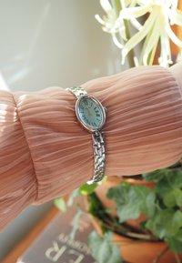 Ceas de mână oval argintiu cu cadran albastru deschis și cifre romane pe o brățară metalică legată, așezat pe o mânecă de țesătură piersică texturată.