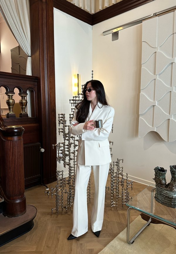 Kvinne i hvit dress og solbriller holder en metallkopp, står i rom med trepanel, moderne skulptur og glassbord med keramisk dekor.