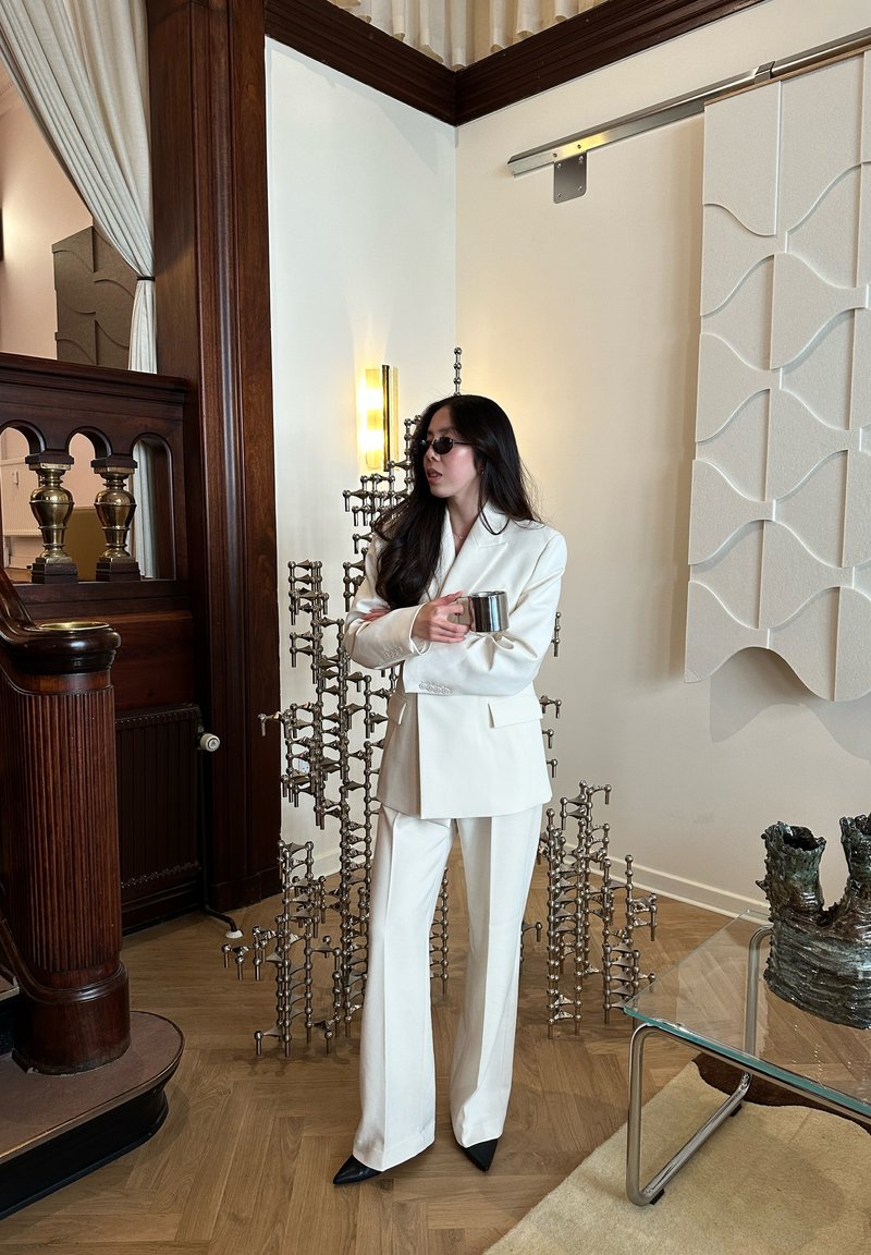 Donna in completo bianco e occhiali da sole tiene una tazza di metallo, in piedi in una stanza con pannelli di legno, scultura moderna e tavolo di vetro con decorazioni in ceramica.