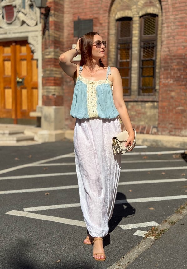 Femme portant des lunettes de soleil, un haut bleu sans manches, un pantalon blanc large et des sandales, tenant une pochette beige, marchant dans une rue ensoleillée près d'un bâtiment en briques.