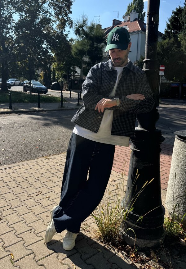 Homme portant une casquette verte, une veste à carreaux, une chemise blanche et un pantalon foncé, appuyé contre un lampadaire noir sur un trottoir urbain ensoleillé.
