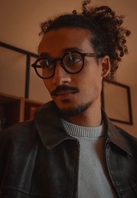 Homme aux cheveux bouclés attachés, portant des lunettes rondes, une veste en cuir et un pull léger, regardant légèrement sur le côté à l'intérieur.