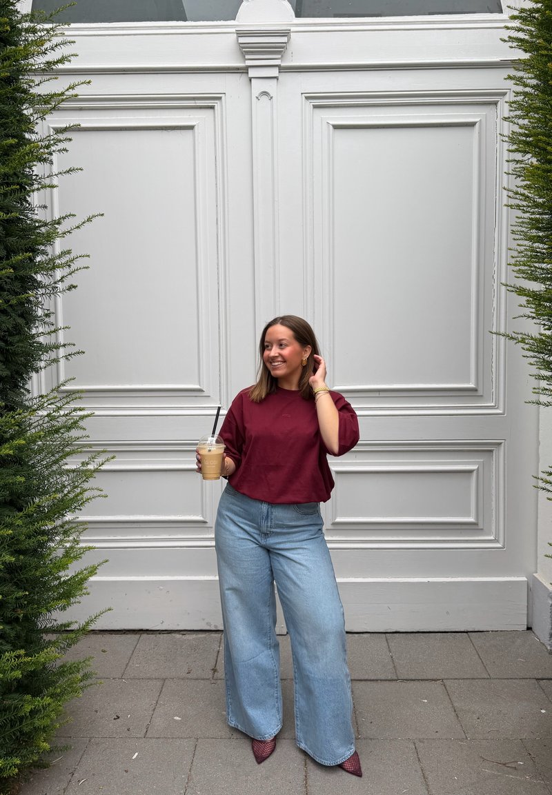 Femme en chemise rouge et jean large bleu tient un café glacé, debout entre deux buissons verts devant de grandes portes blanches doubles.