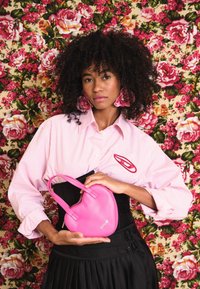 Bolsa em forma de coração rosa, feita de material suave, segurada por uma pessoa vestindo uma camisa rosa de riscas e uma saia preta, com um fundo floral.