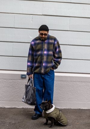 Chaqueta de forro polar con patrón de cuadros en azul, verde y rosa; jeans azul de corte holgado; sosteniendo una bolsa transparente. Perro marrón con un abrigo verde forrado en blanco.