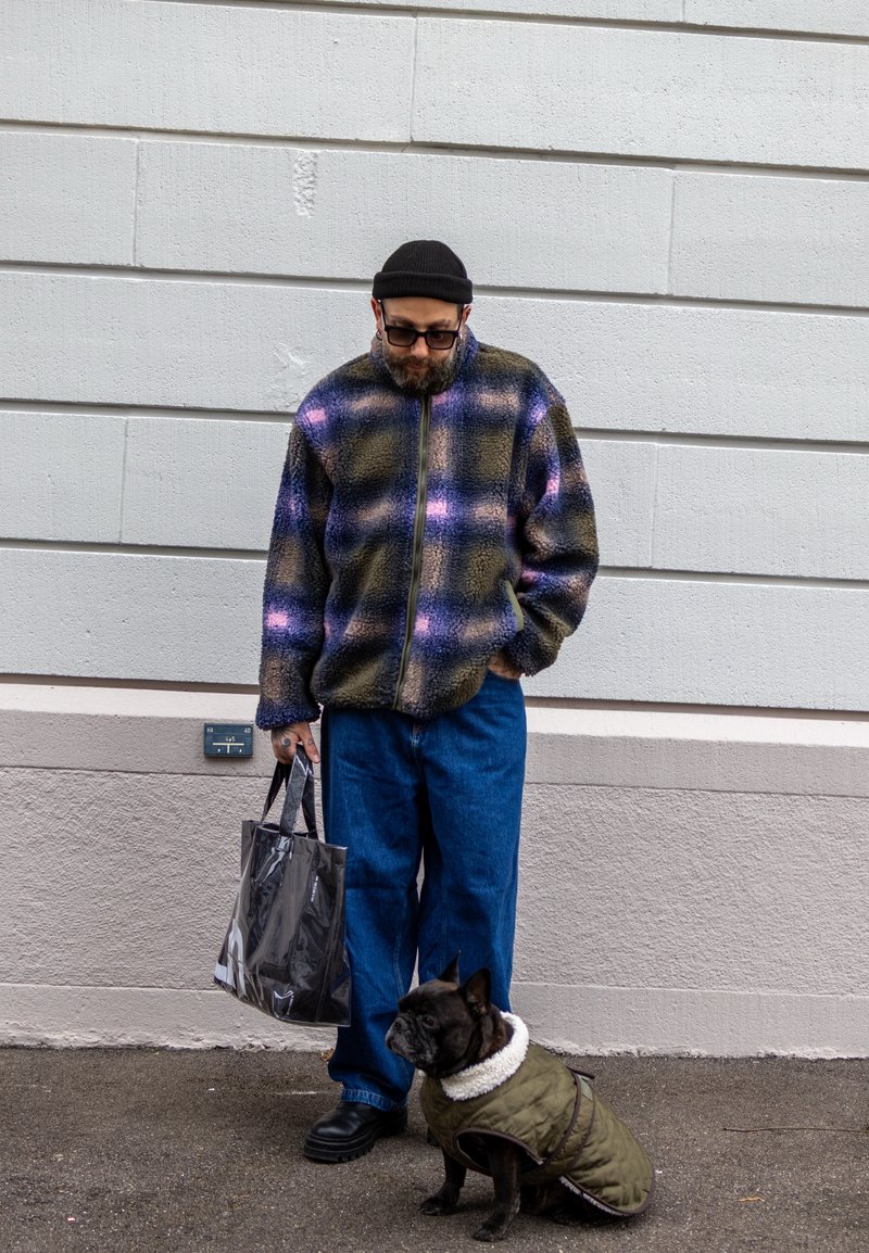 Veste polaire à carreaux bleus, verts et roses ; jean bleu coupe ample ; tenant un sac transparent. Chien brun portant un manteau vert avec doublure blanche.