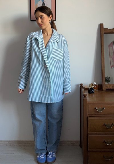 Camisa de rayas azul claro y blanca de corte oversized con un bolsillo parche, combinada con jeans sueltos azules y zapatillas azules. Mueble de madera en el fondo.