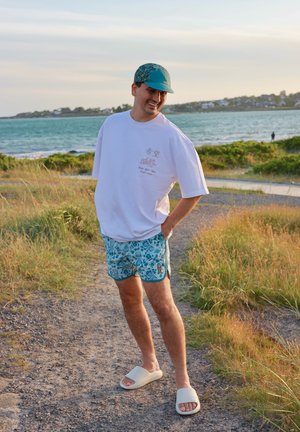 Camiseta blanca de gran tamaño con un gráfico, pantalones cortos de flores turquoise, sandalias ligeras de tipo deslizar, de pie en un camino de grava.