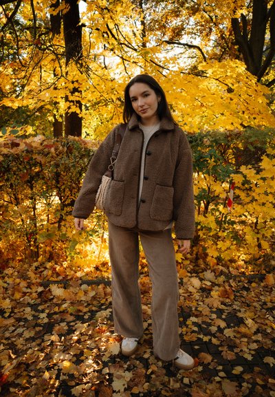 Joven mujer de pie en un camino cubierto de hojas, vistiendo una chaqueta de forro polar marrón, pantalones de pierna ancha beige y zapatillas blancas, con árboles de otoño al fondo.